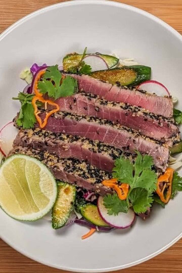 Sesame crusted ahi tuna on a bed of cabbage slaw, carrots, radishes, and zucchini with soy-sesame dressing and garnished with fresh cut lime, cilantro leaves and habanero slices on a wide rim bowl.