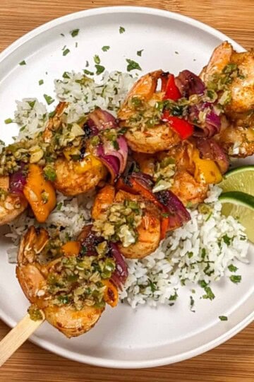 two Grilled Shrimp skewers with Garlic Jalapeno Butter and Cilantro Lime Rice on a flat round plate garnished with lime wedges and chopped cilantro.