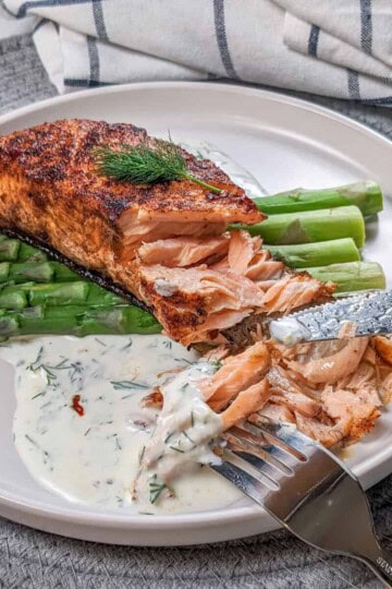 smoked paprika Calabrian Pepper marinated pan-seared Salmon with Creamy Lemon Dill Sauce and jumbo asparagus on a round white plate being eaten with a fork and knife.