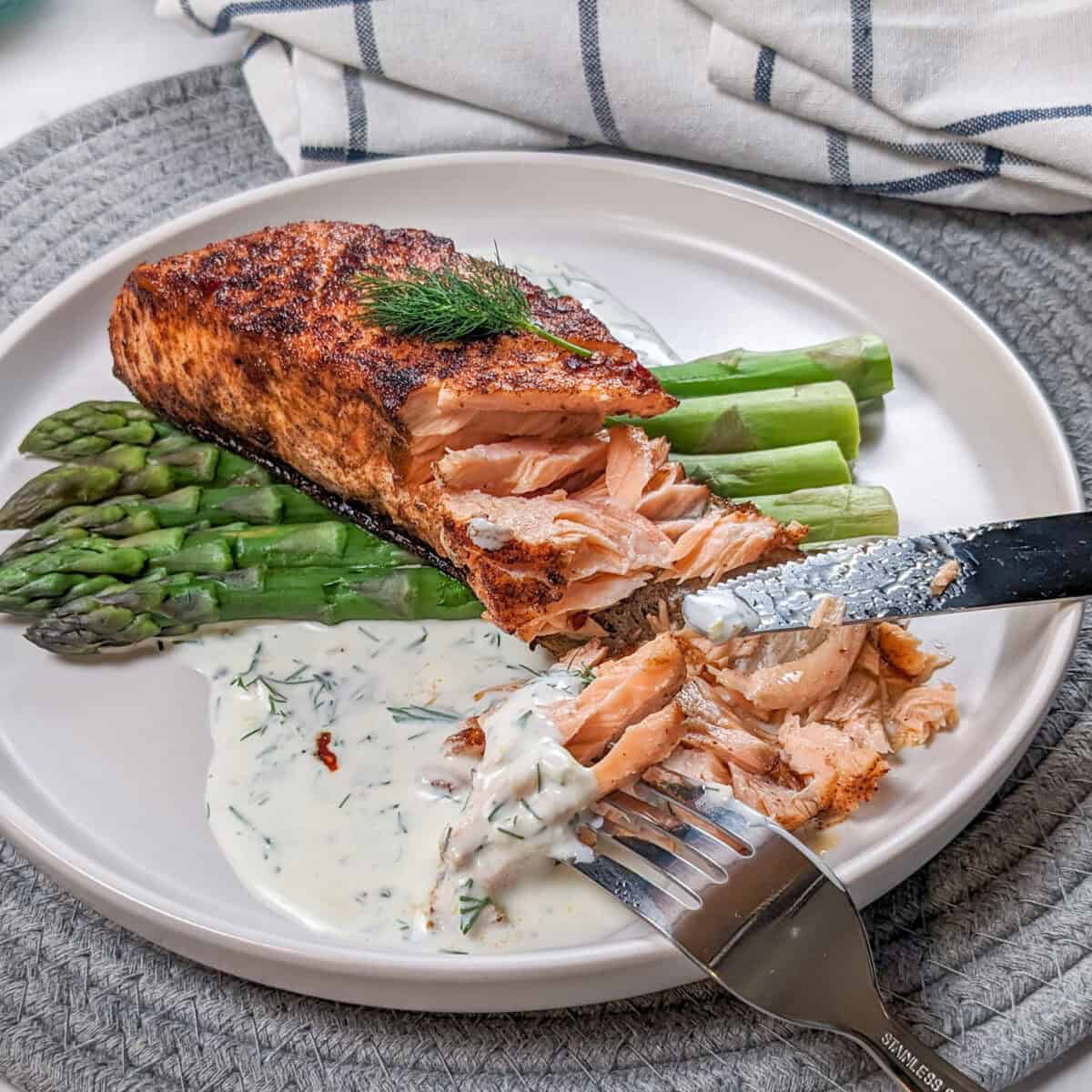 smoked paprika Calabrian Pepper marinated pan-seared Salmon with Creamy Lemon Dill Sauce and jumbo asparagus on a round white plate being eaten with a fork tested on the cooked fish and a knife.