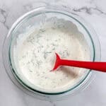 creamy lemon dill sauce in a glass pyrex mixing bowl with a red silicone spoon sitting in it.