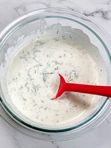 creamy lemon dill sauce in a glass pyrex mixing bowl with a red silicone spoon sitting in it.