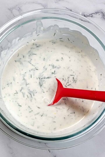 creamy lemon dill sauce in a glass pyrex mixing bowl with a red silicone spoon sitting in it.