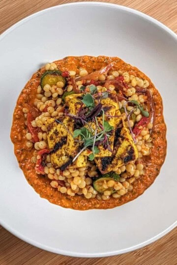 grilled marinated tofu on top of a warm roasted vegetable couscous salad on spicy basil romesco sauce in a wide rim bowl topped with micro greens.
