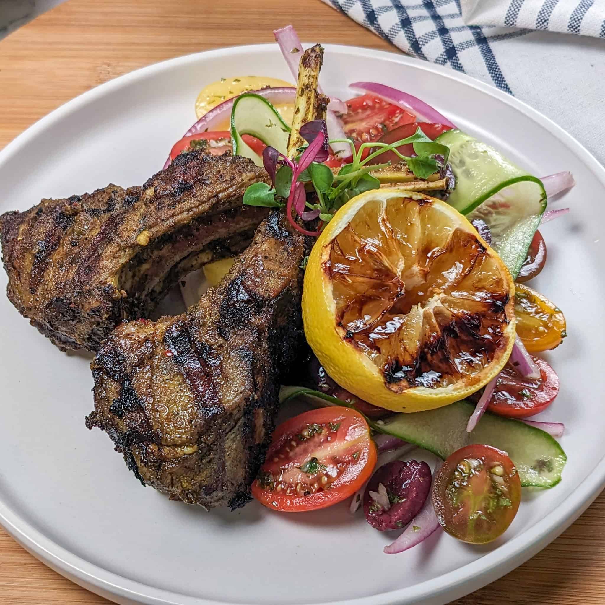 Grilled Zhug Ras el Hanout Lamb Chops and Tomato, cucumber, onion Salad served with a lemon-oregano dressing served with a grilled lemon.