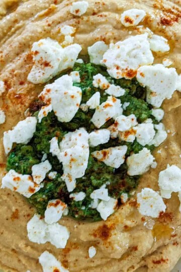 close up of chunky hummus topped with zhug in the center and sprinkled with crumbled feta cheese and smoked paprika and drizzled with olive oil.