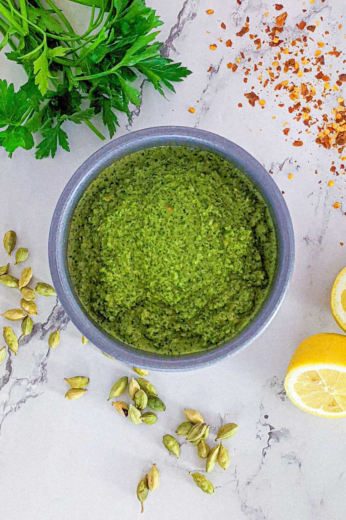 Zhug sauce in a bowl surrounded by lemons, chili pepper flakes, fresh parsley and cardamom pods.