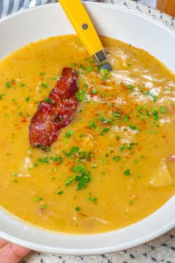 wide rim bowl filled with Creamy spicy bacon potato leek chowder that's garnished with a half of slice of crispy bacon and chopped parsley then dusted with smoked paprika held in hand.