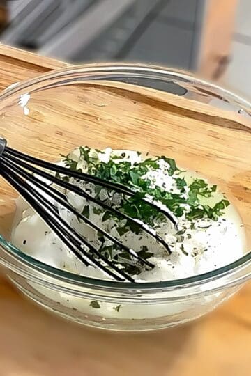 lemon greek yogurt with chopped parsley on top in a glass mixing bowl with a silicone whisk.