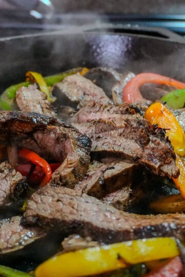 sliced jerk seasoned steak fajitas withs sliced tri color sweet bell peppers and onions in a steaming cast iron skillet.