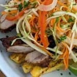 loaded plantain fries in a round dish with tall sides topped with sliced seasoned chicken thighs and pikliz (a hot pepper cabbage slaw).