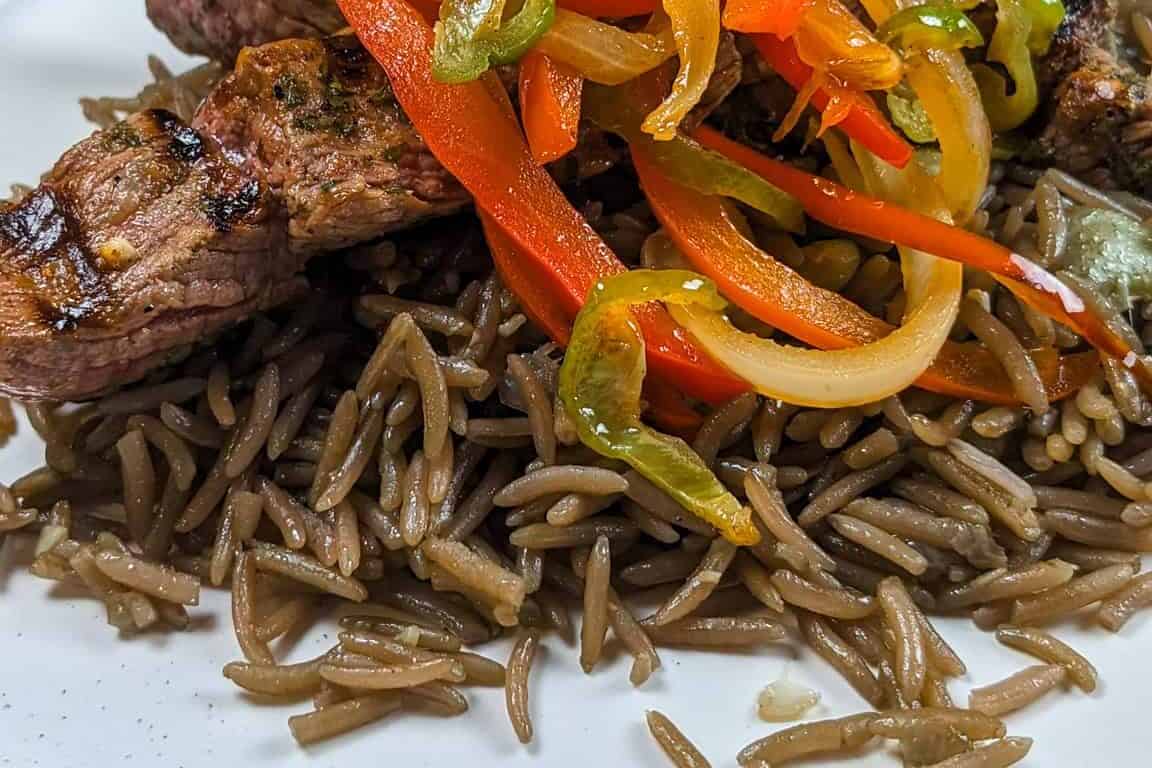 side view of the Grilled beef skewers with black mushroom rice on a flat round plate with more escovitch-style vegetables in the background.