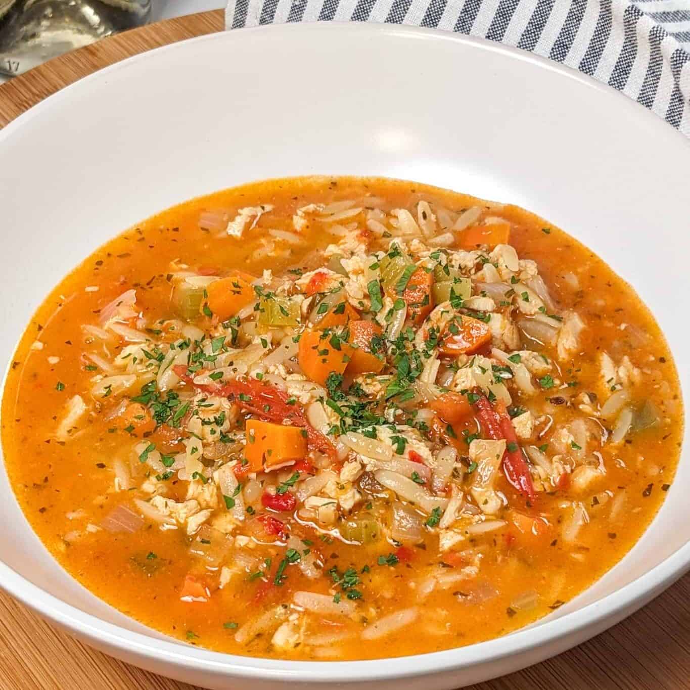 Lemon Calabrian Chili Chicken Orzo Soup in a wide rim bowl on a lazy susan with a kitchen towel and white wine bottle in the background.