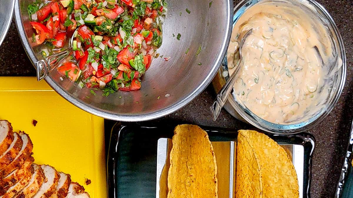cholula green chli sour cream in a glass bowl with a spoon surrounded by taco shells, sliced seasoned chicken and pico de gallo.
