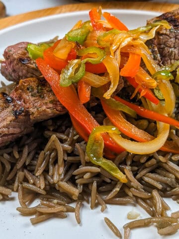 side view of the Grilled beef skewers with black mushroom rice on a flat round plate with more escovitch-style vegetables in the background.