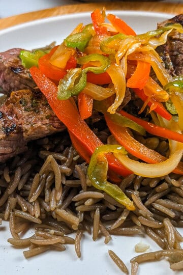 side view of the Grilled beef skewers with black mushroom rice on a flat round plate with more escovitch-style vegetables in the background.