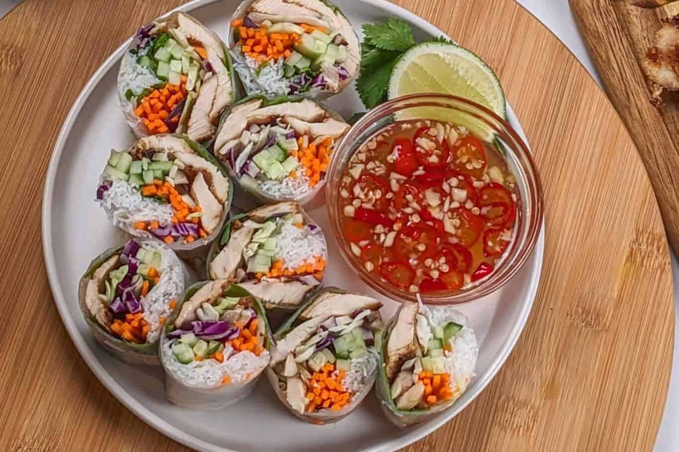 sliced lemongrass chicken, cabbage, rice vermicelli spring roll with nouc cham sauce on side all on a round flat plate surrounded by glass container of shredded tri-color slaw, and other ingredients.