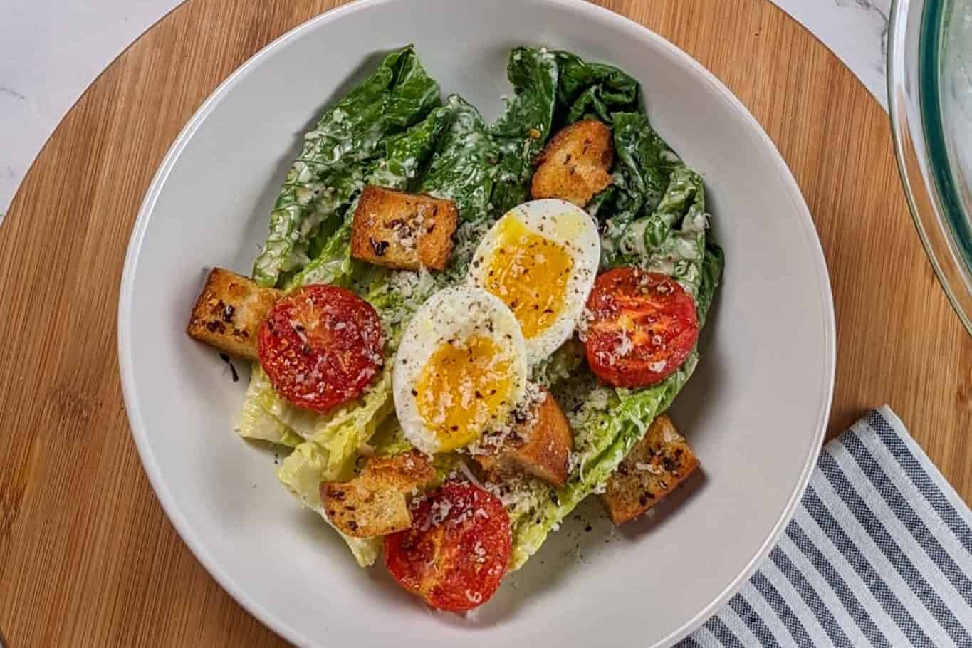 Caesar Salad with soft-boiled Egg, roasted campari Tomatoes and Chili-Infused Croutons garnished with freshly grated parmesan cheese and cracked black pepper in a wide rim bowl surrounded by a quarter sheet pan and rack with more roasted halved tomatoes, another sheet pan of croutons, parmesan wedge and a microplane.
