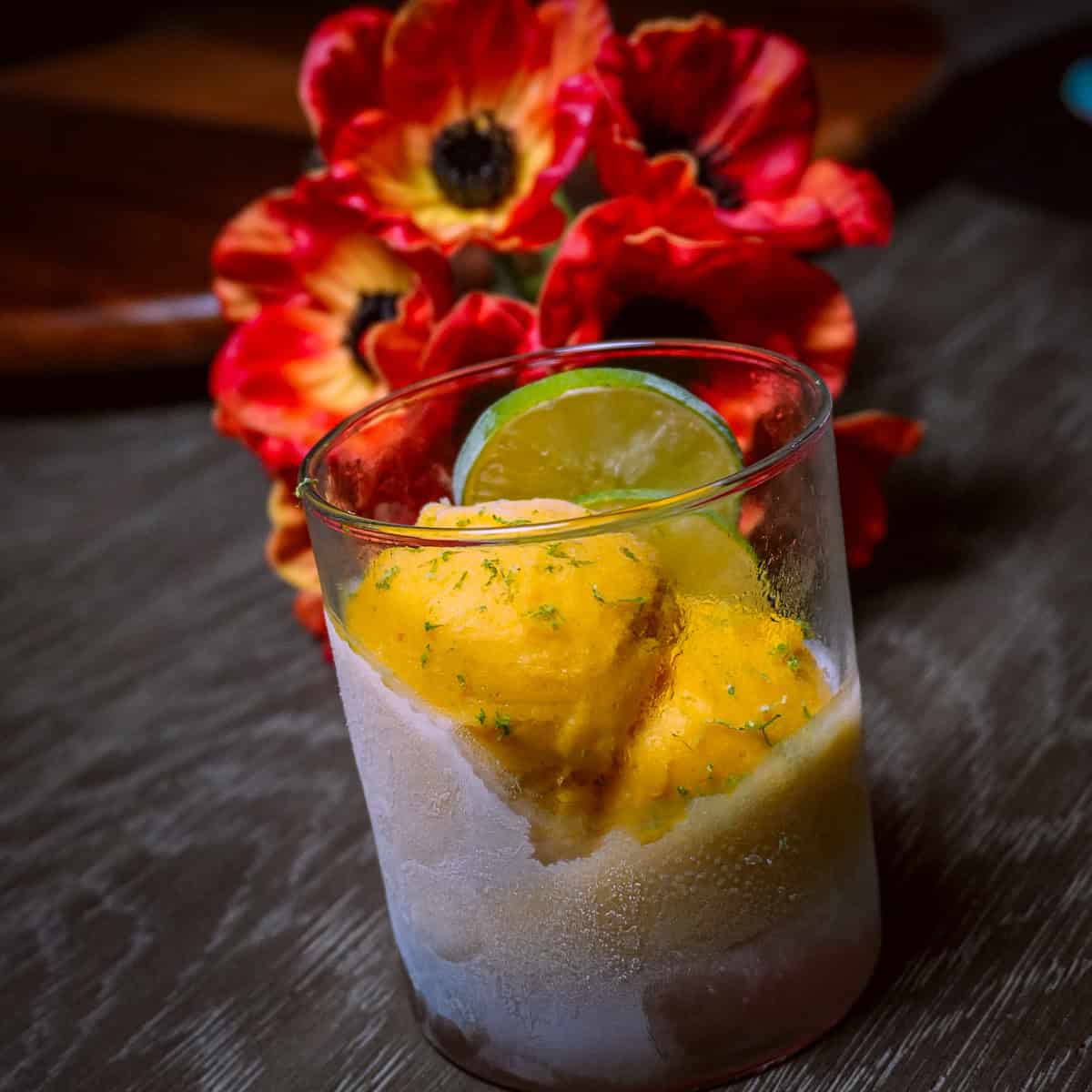 scooped mango coconut felato gelato in glass cup garnished with lime slices and lime zest with poppy flowers in the background on a dining table.