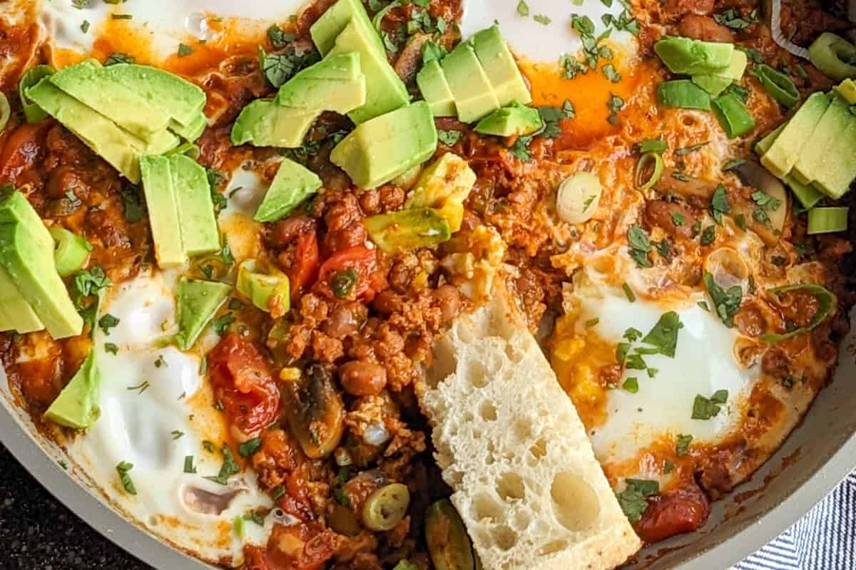 close up of of simmered poached eggs in a skillet with crumbled soy chorizo, seasoned pinto beans, diced fresh avocados and sliced ciabatta bread.