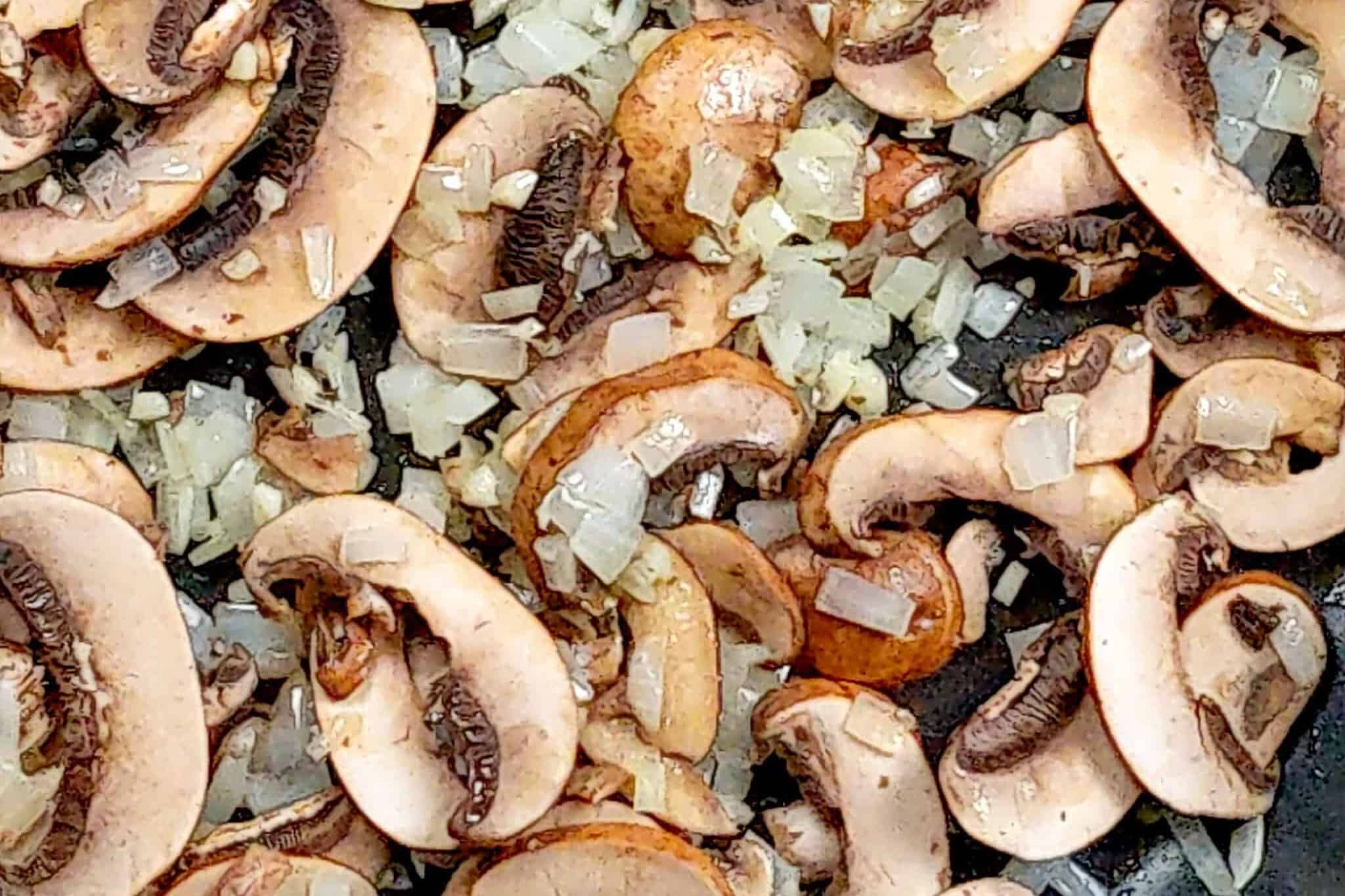 sautéed onions and garlic with raw sliced mushrooms in a frying pan.