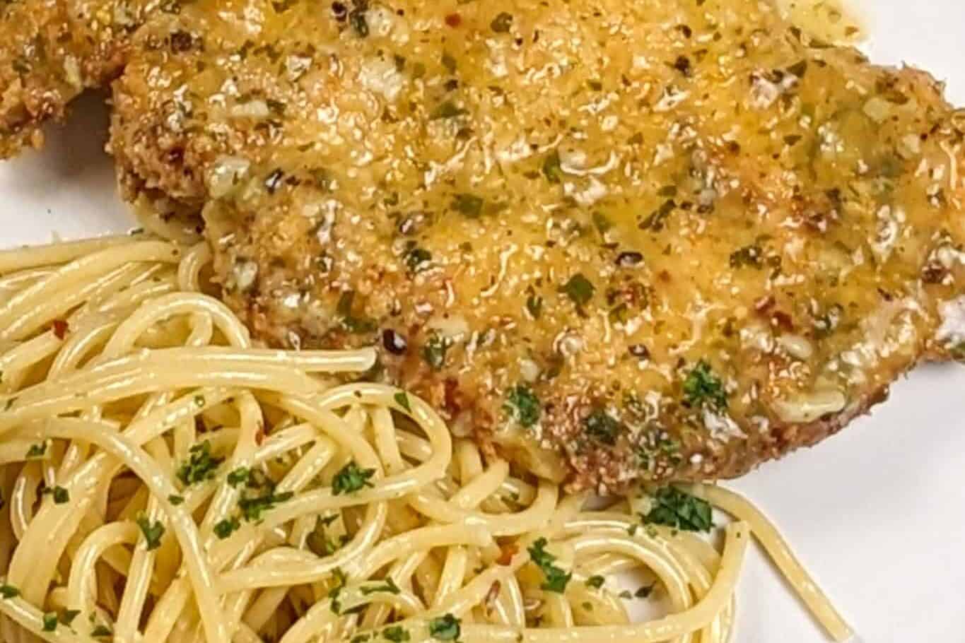 wet lemon pepper chicken cutlet on a white round plate next to olive oil and grated cheese tossed spaghetti dusted with chopped fresh parsley