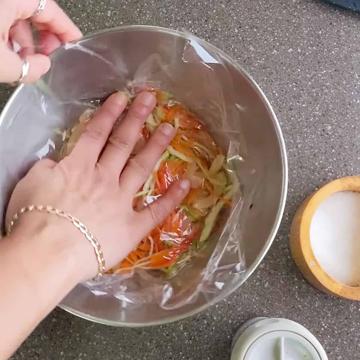 plastic wrap being gently pressed on top of the pikliz spicy slaw mixture.