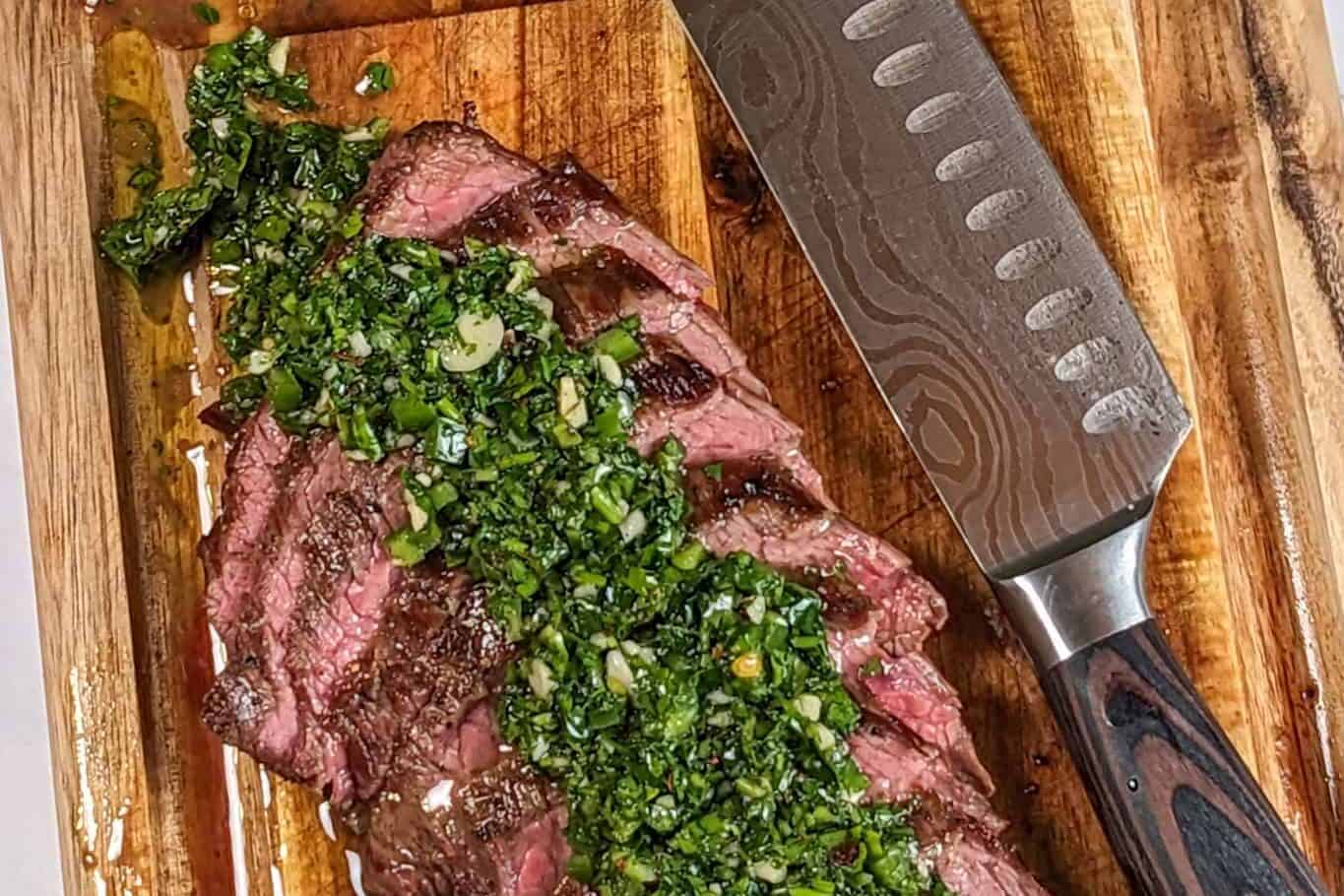 medium rare thinly sliced pan-grilled flank steak topped with spicy chimichurri sauce cascading on a mini wooden cutting board with a knife laying next to it.