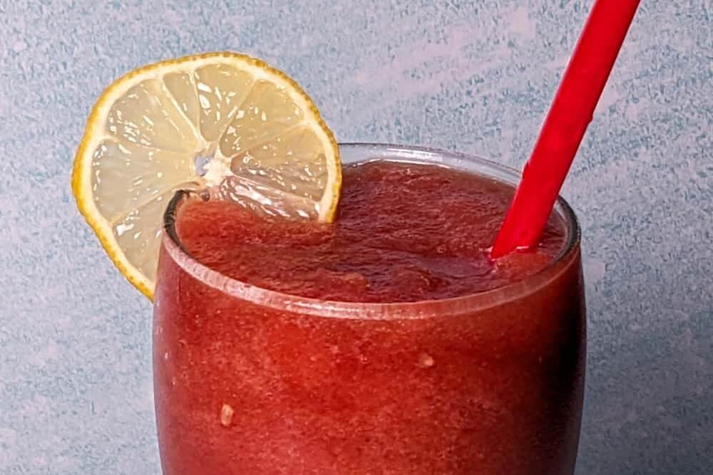 frozen watermelon lemonade in a tall glass with a red straw and garnished with a fresh lemon wheel.