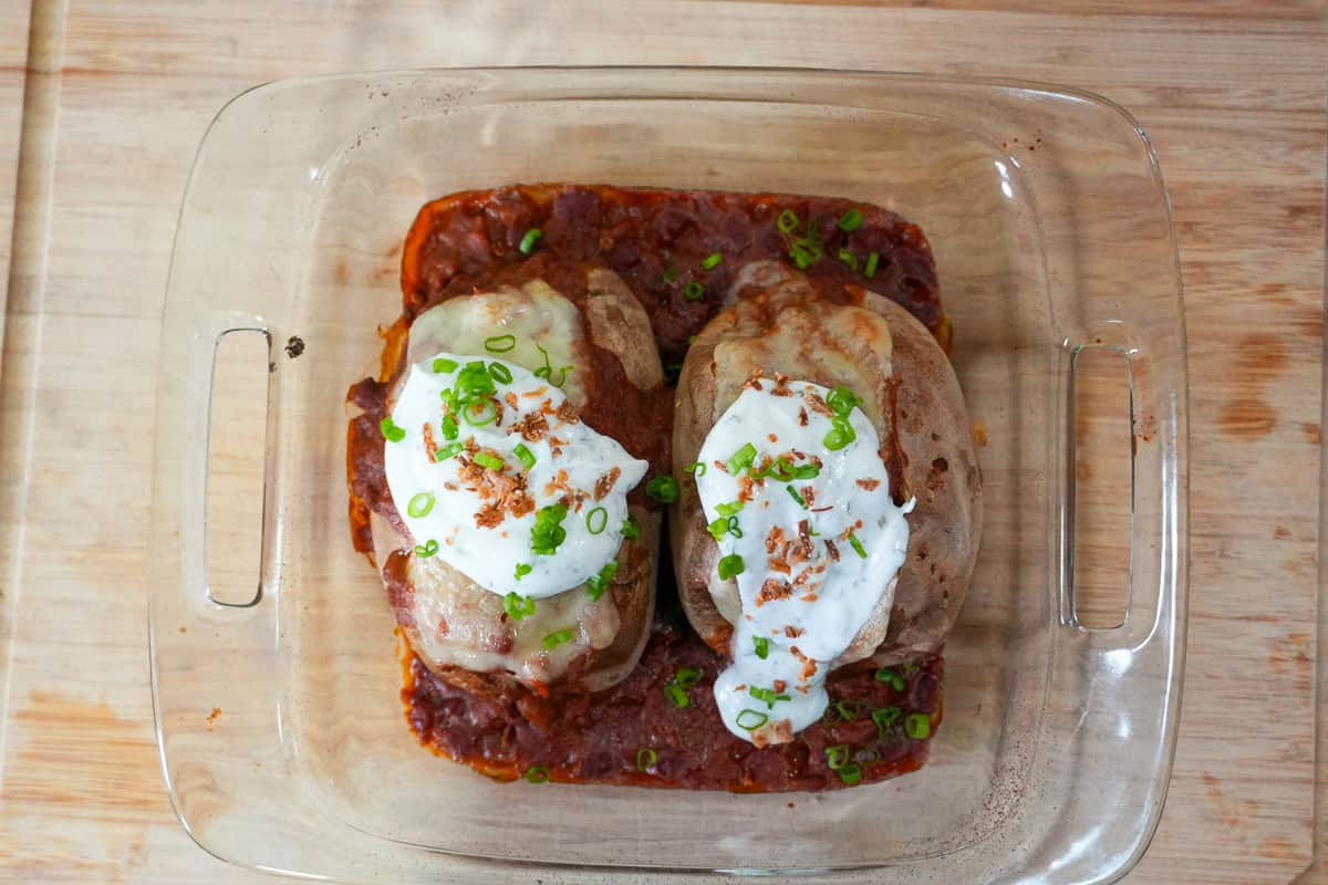 baked potatoes and chili with melted swiss cheese in a baking dish topped with seasoned sour cream and garnishes.