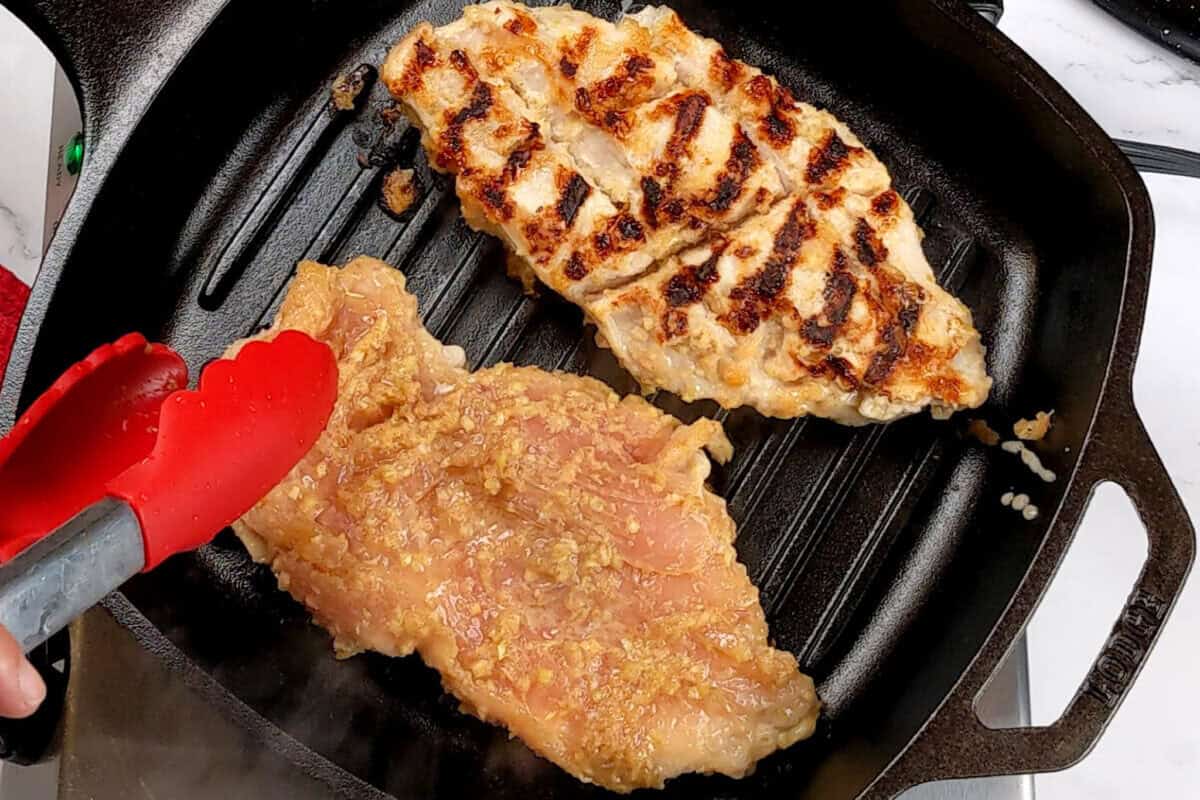 two grilled lemongrass chicken breast with grill marks on one and another about to be flipped with a kitchenaid silicone spatula in a grill pan.