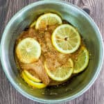 bowl of sliced chicken breast, marinating in seasonings and sliced lemons in a mixing bowl.