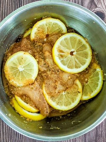 bowl of sliced chicken breast, marinating in seasonings and sliced lemons in a mixing bowl.