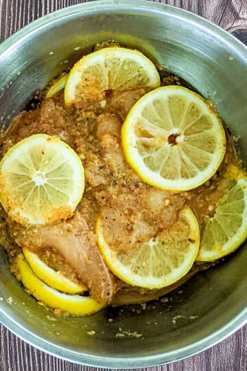 bowl of sliced chicken breast, marinating in seasonings and sliced lemons in a mixing bowl.