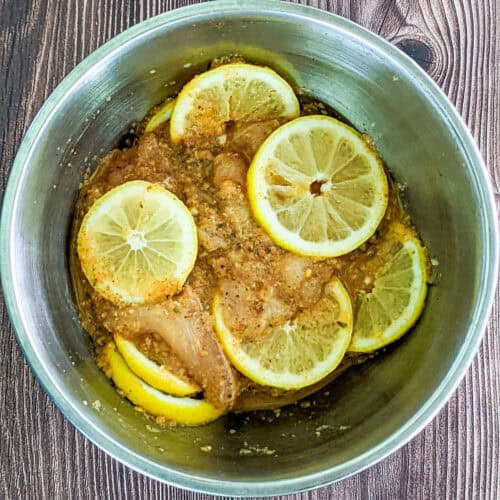bowl of sliced chicken breast, marinating in seasonings and sliced lemons in a mixing bowl.