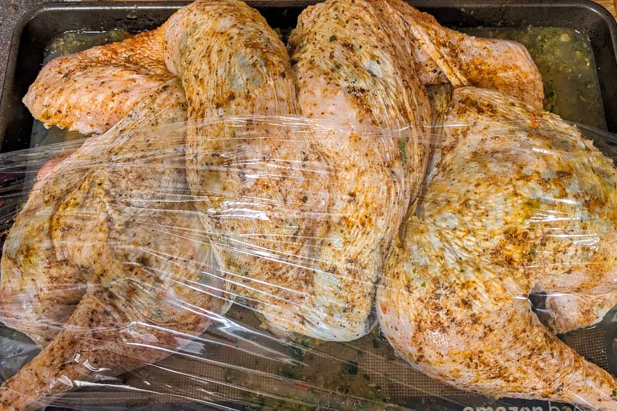 haitian style seasoned spatchcock turkey on a sheet tray covered in plastic wrap.
