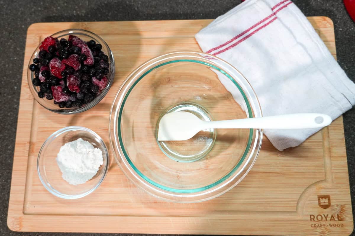 cutting board topped with three glass bowls with frozen berries, flour, and spatula with a kitchen towel.
