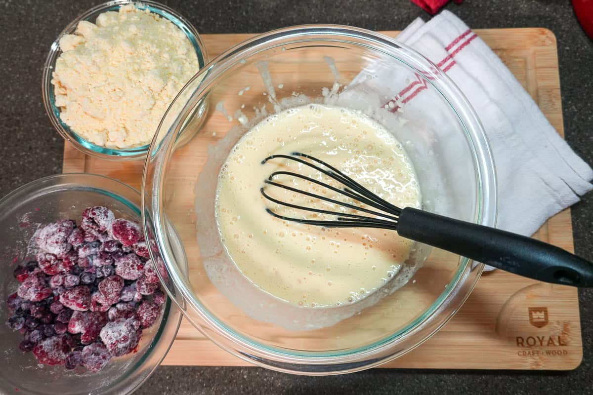 wet ingredients mixed in the mixing bowl with a whisk with two bowls filled with jiffy corn muffin mix and frozen blueberries and raspberries tossed in flour.