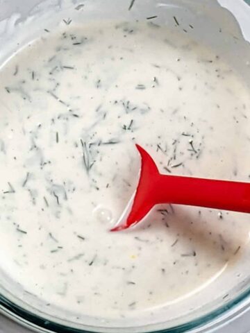 creamy lemon dill sauce in a glass pyrex mixing bowl with a red silicone spoon sitting in it.