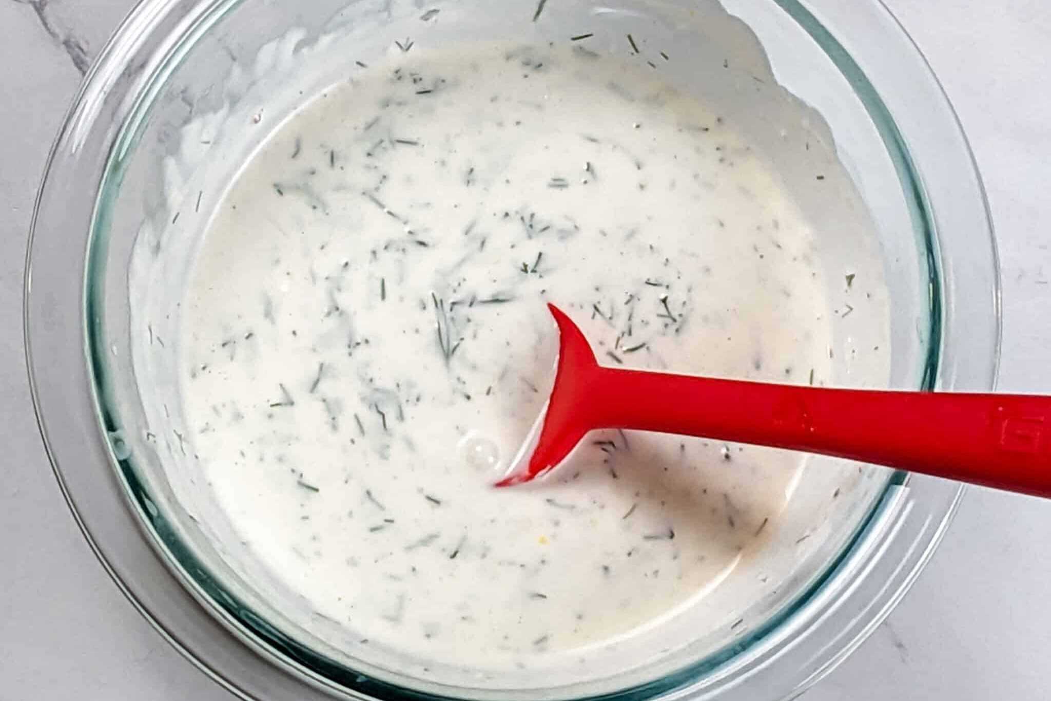 creamy lemon dill sauce in a glass pyrex mixing bowl with a red silicone spoon sitting in it.