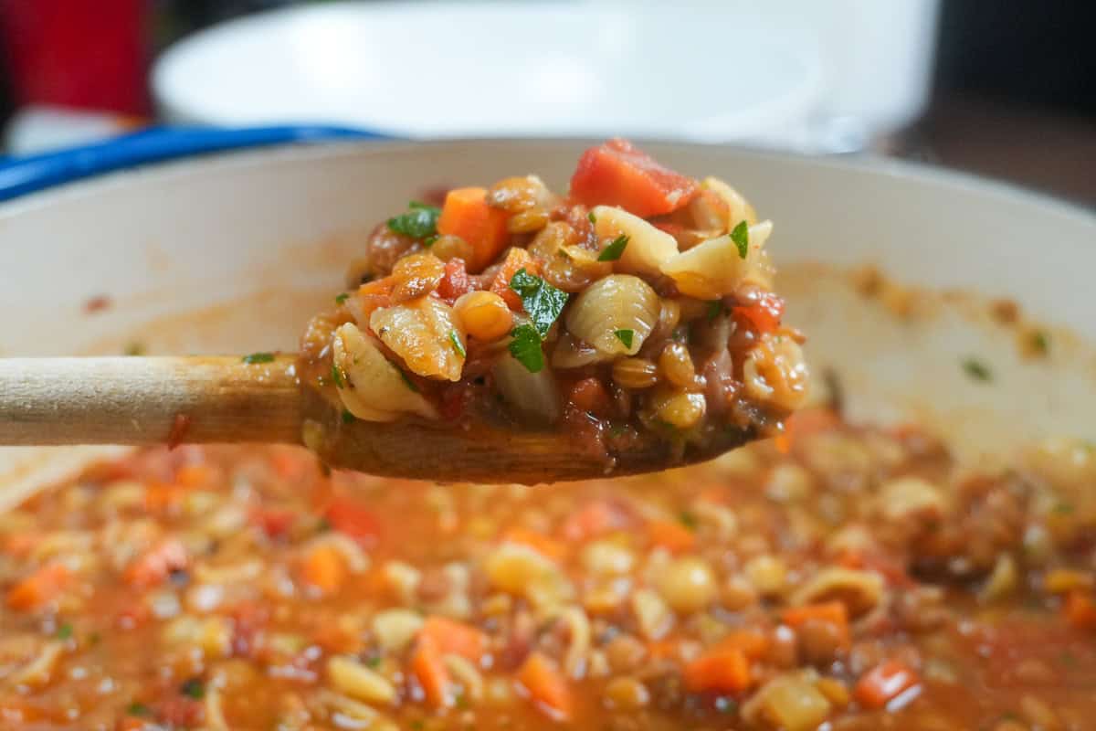 close up of the Pasta and Lentil Soup Recipe on a wooden spoon over a dutch oven fill with the soup.