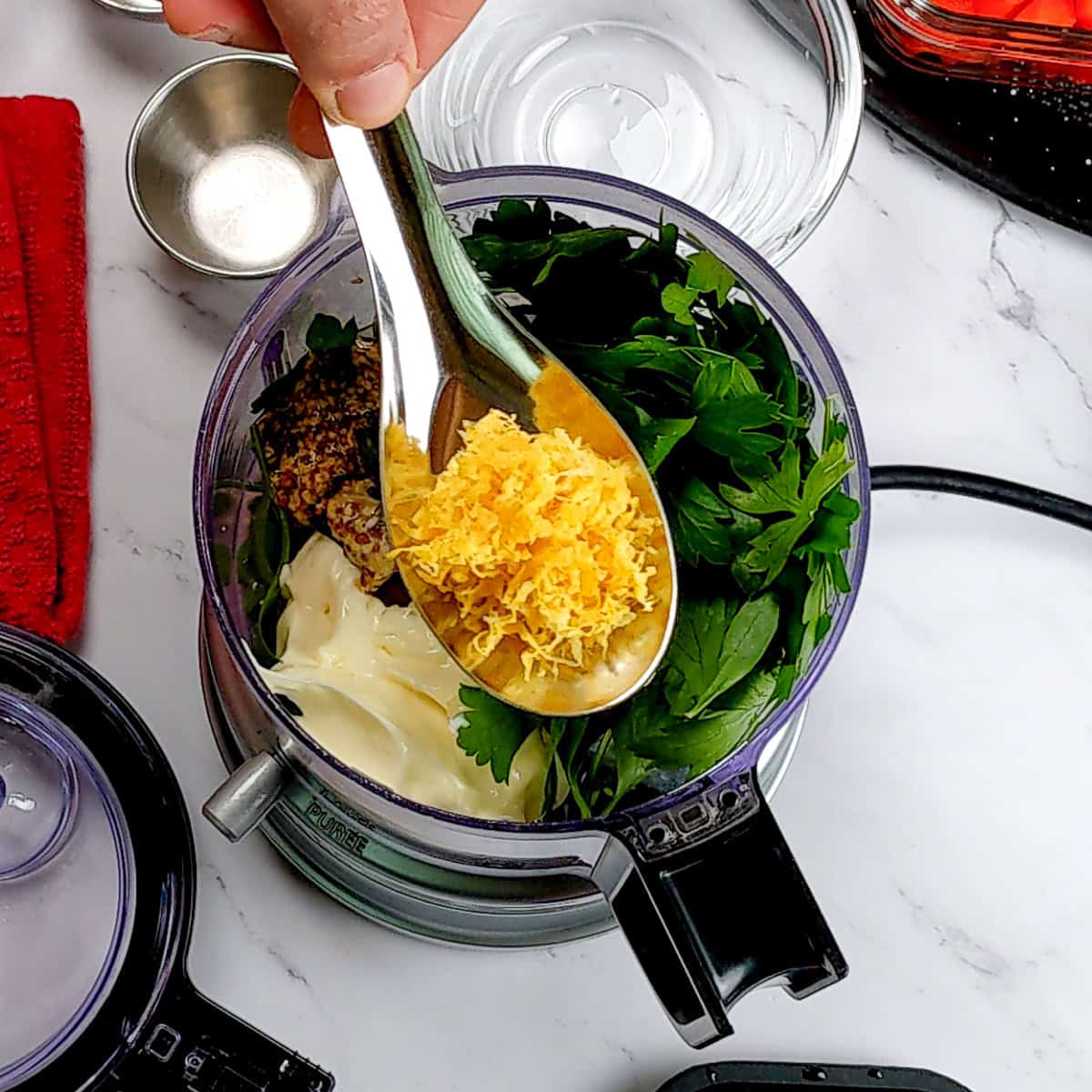whole ingredients in the food chopper with the lemon zest on a spoon on top.