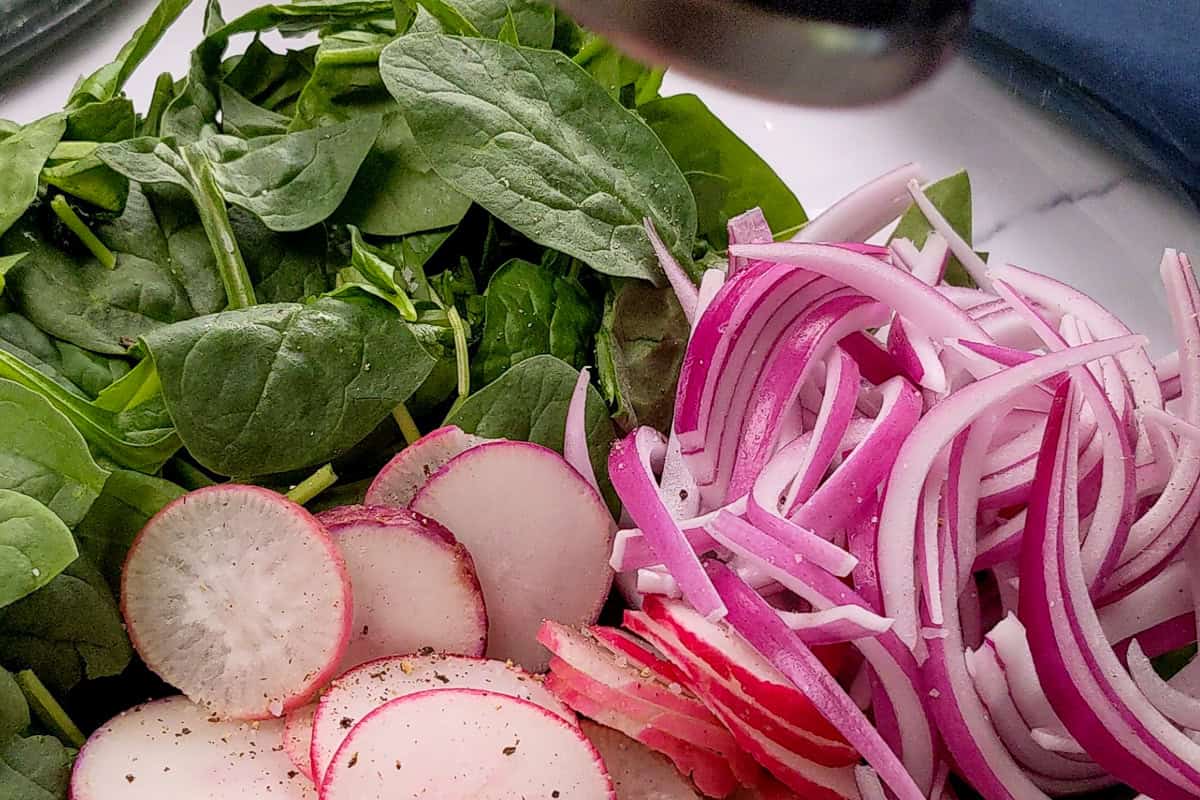 Spring produce of sliced radishes, baby spinach and onions in a glass bowl.