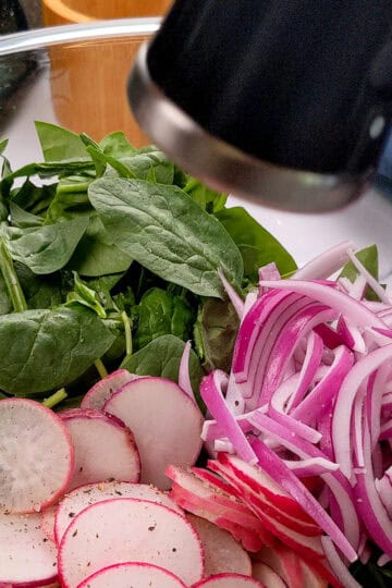 Spring produce of sliced radishes, baby spinach and onions in a glass bowl.