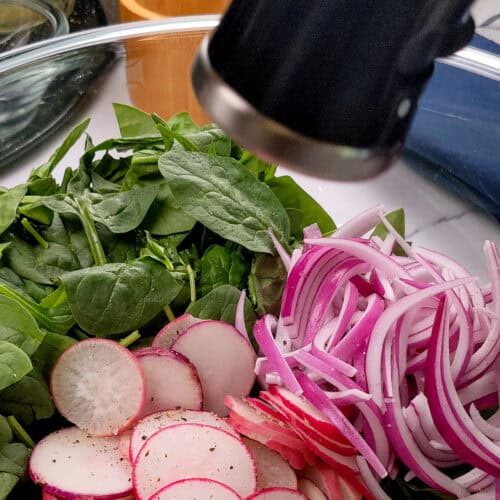 Spring produce of sliced radishes, baby spinach and onions in a glass bowl.