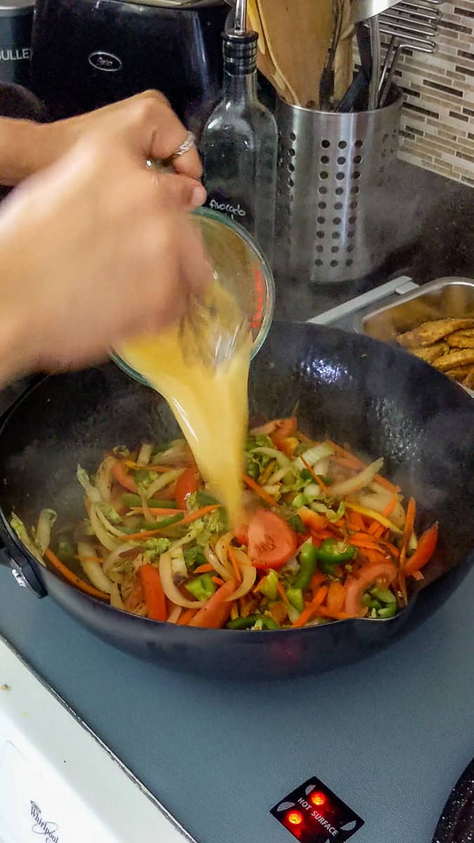 sauce from the glass measuring cup being poured over the stir-fried vegetables for the jamaican chicken curry.