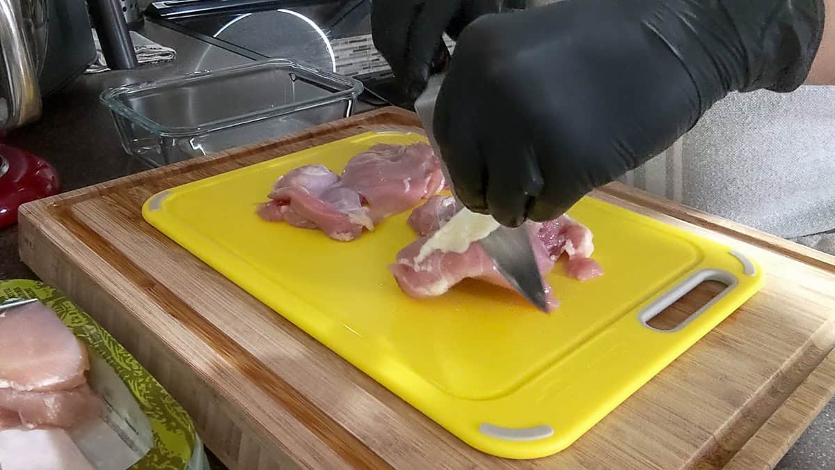 using fingers to pull up fat on chicken thigh with knife in other hand to trim the fat away from self.