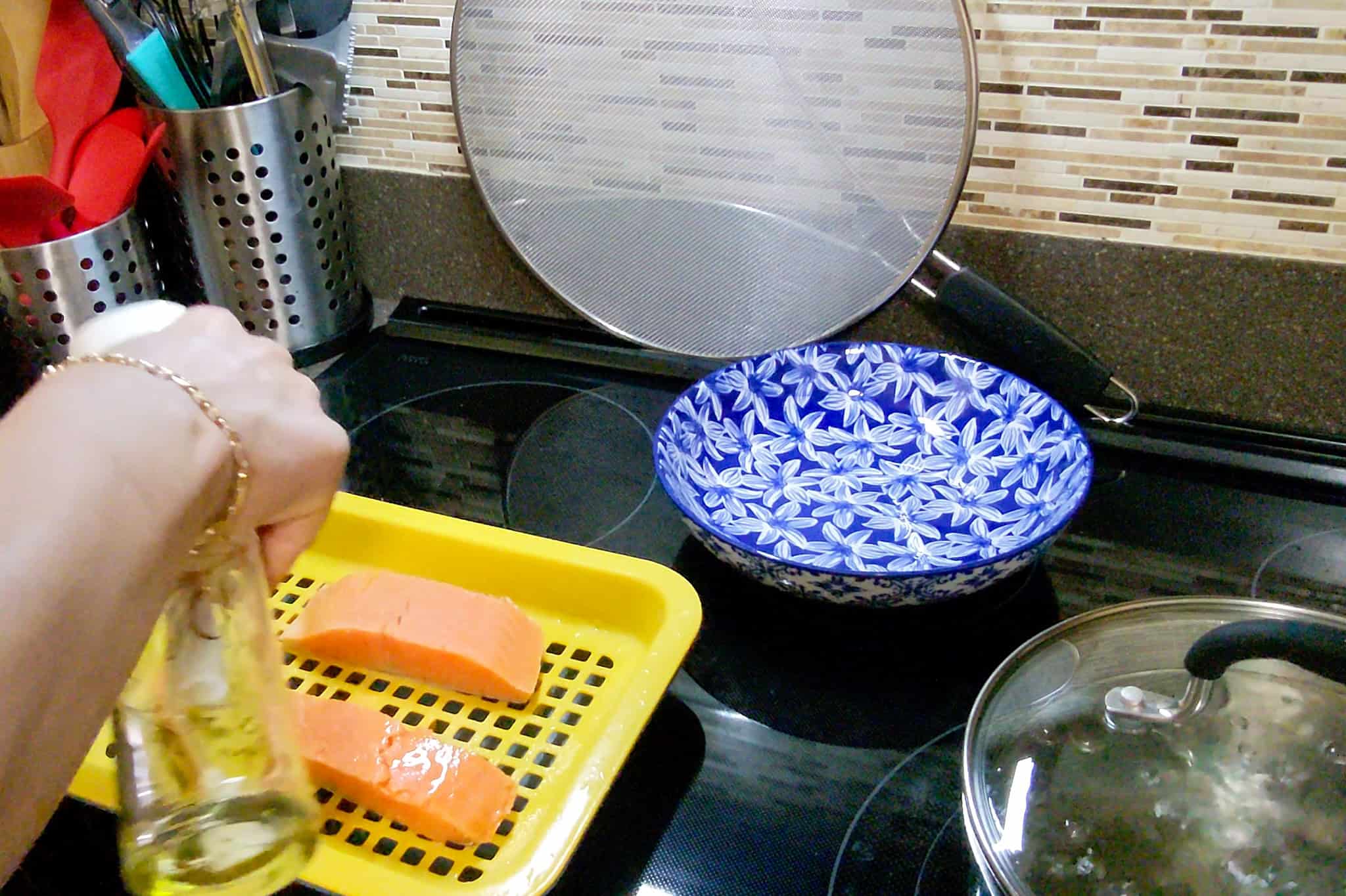 Chef Maika oil spraying salmon fillets on a kitchen stove with utensils and cookware.