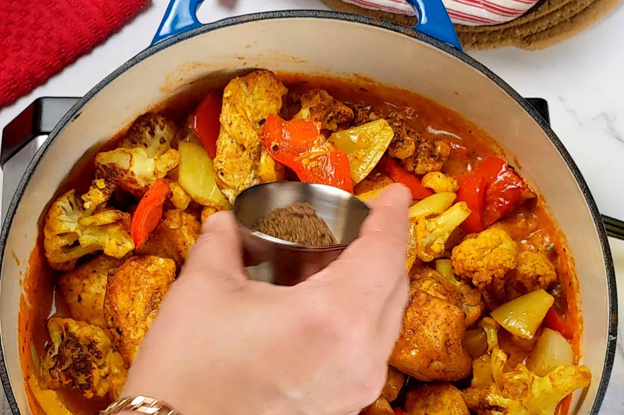 garam masala in a stainless steel condiment cup being poured into the chicken vegetable curry in the dutch oven.