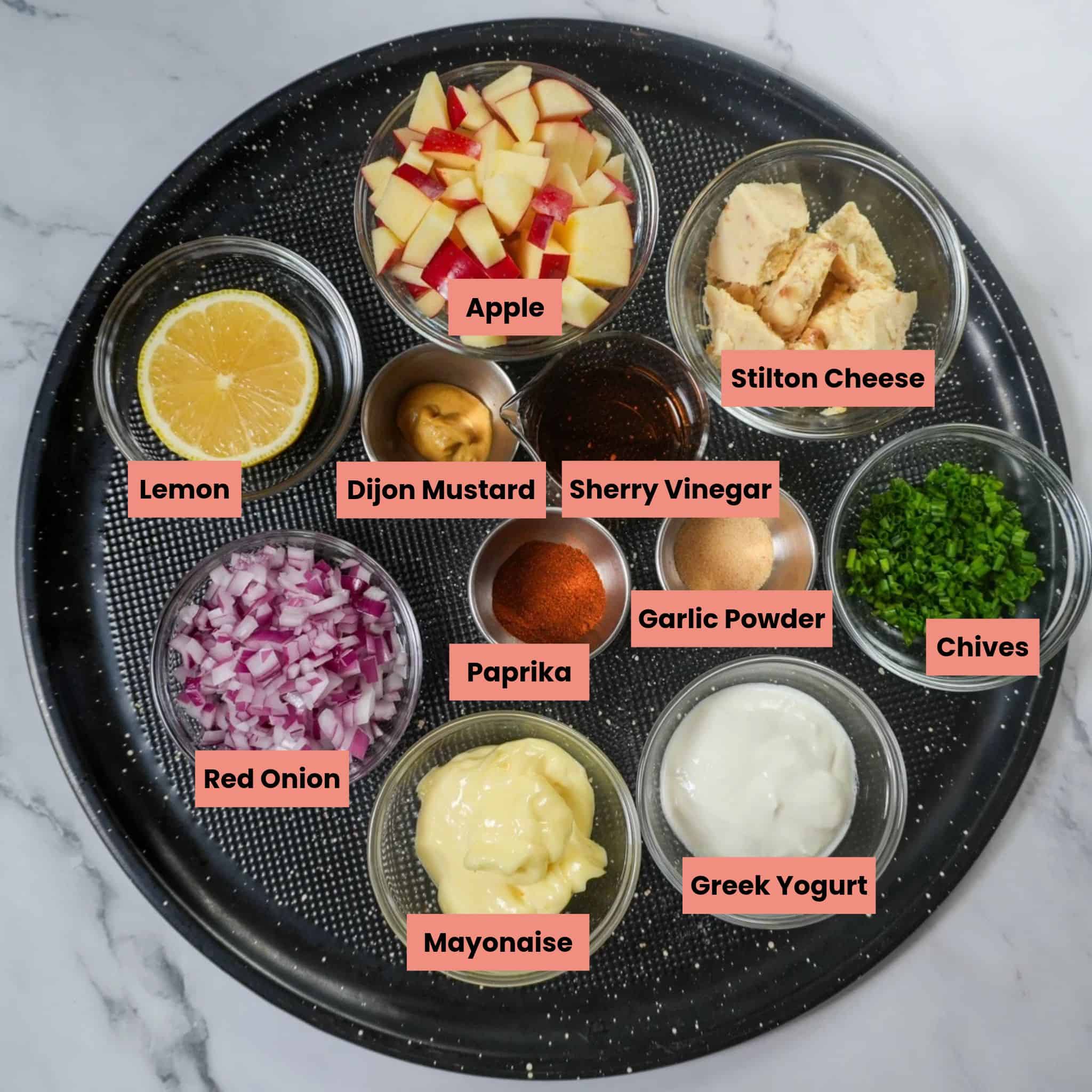 Chicken breasts, fresh apple, red onion, chives, Stilton cheese, and creamy dressing ingredients arranged on a counter on a round tray.